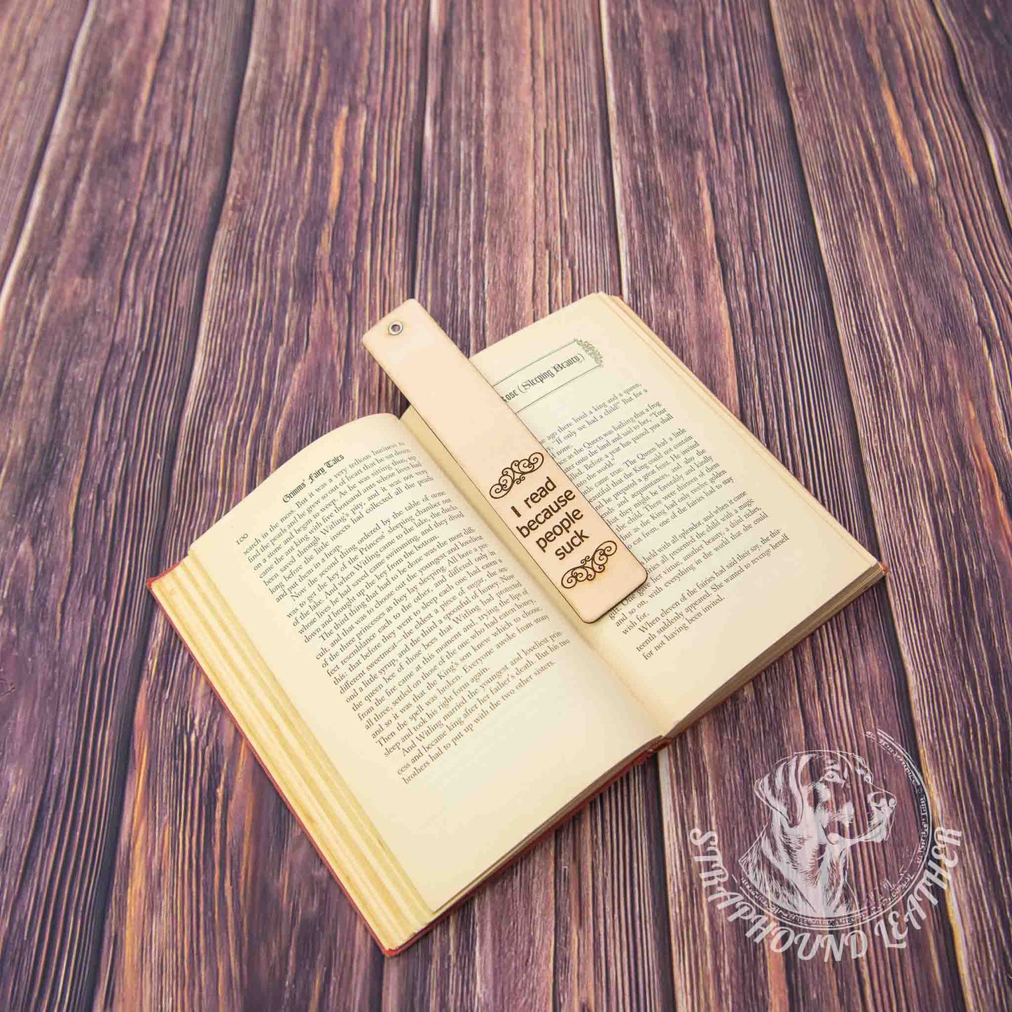 leather bookmark with a scoll-y design and "I read because people suck" laying on an open book on a woodgrain background.
