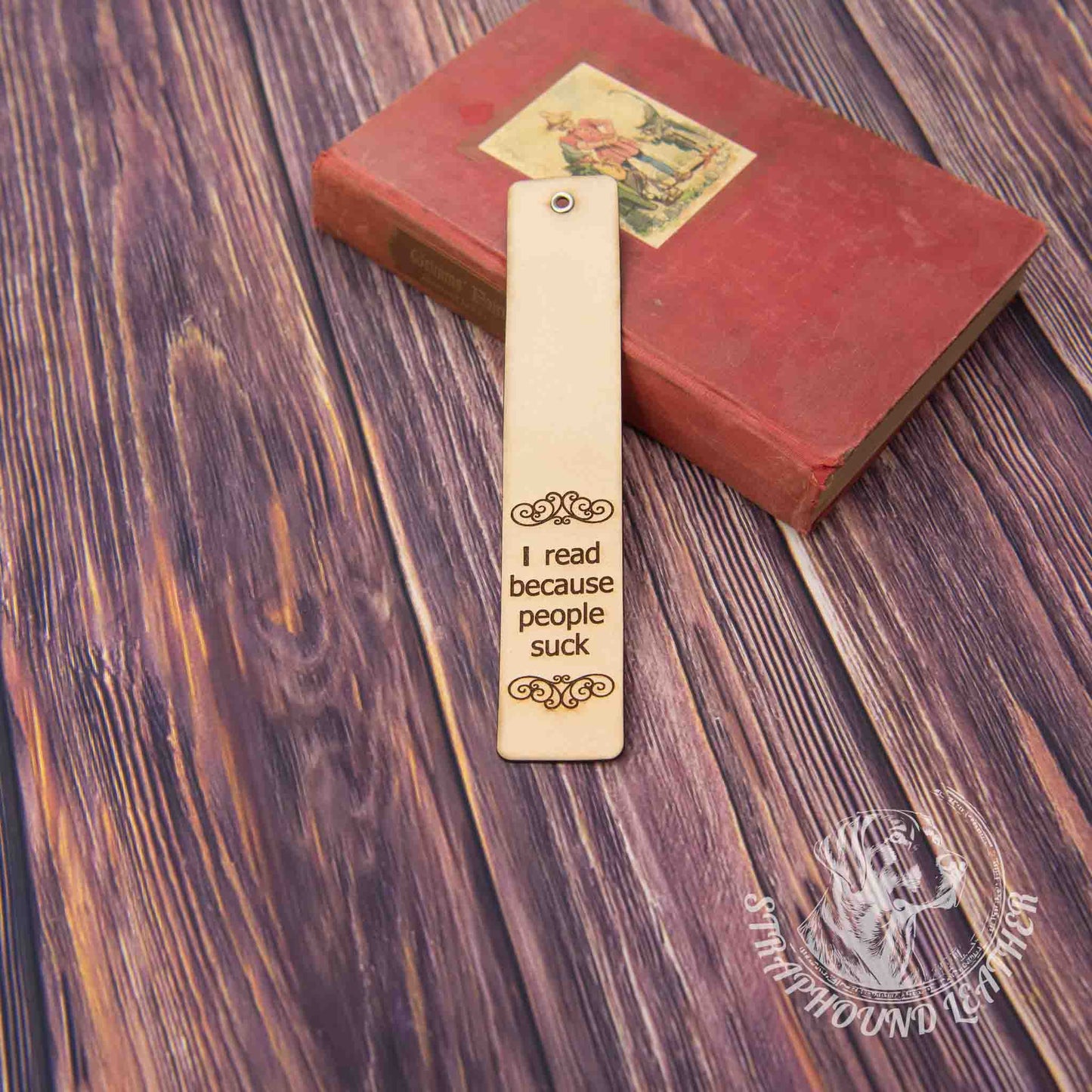 leather bookmark with a scoll-y design and "I read because people suck" leaning against a book on a woodgrain background.
