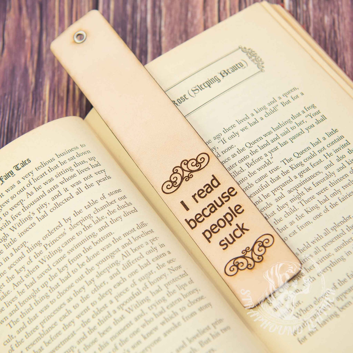 leather bookmark with a scoll-y design and "I read because people suck" laying on an open book on a woodgrain background.
