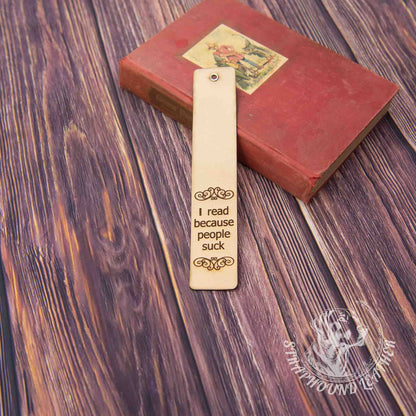 leather bookmark with a scoll-y design and "I read because people suck" leaning against a book on a woodgrain background.