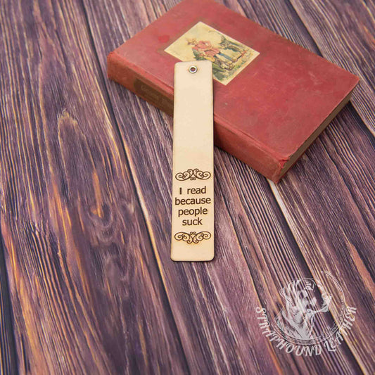 leather bookmark with a scoll-y design and "I read because people suck" leaning against a book on a woodgrain background.